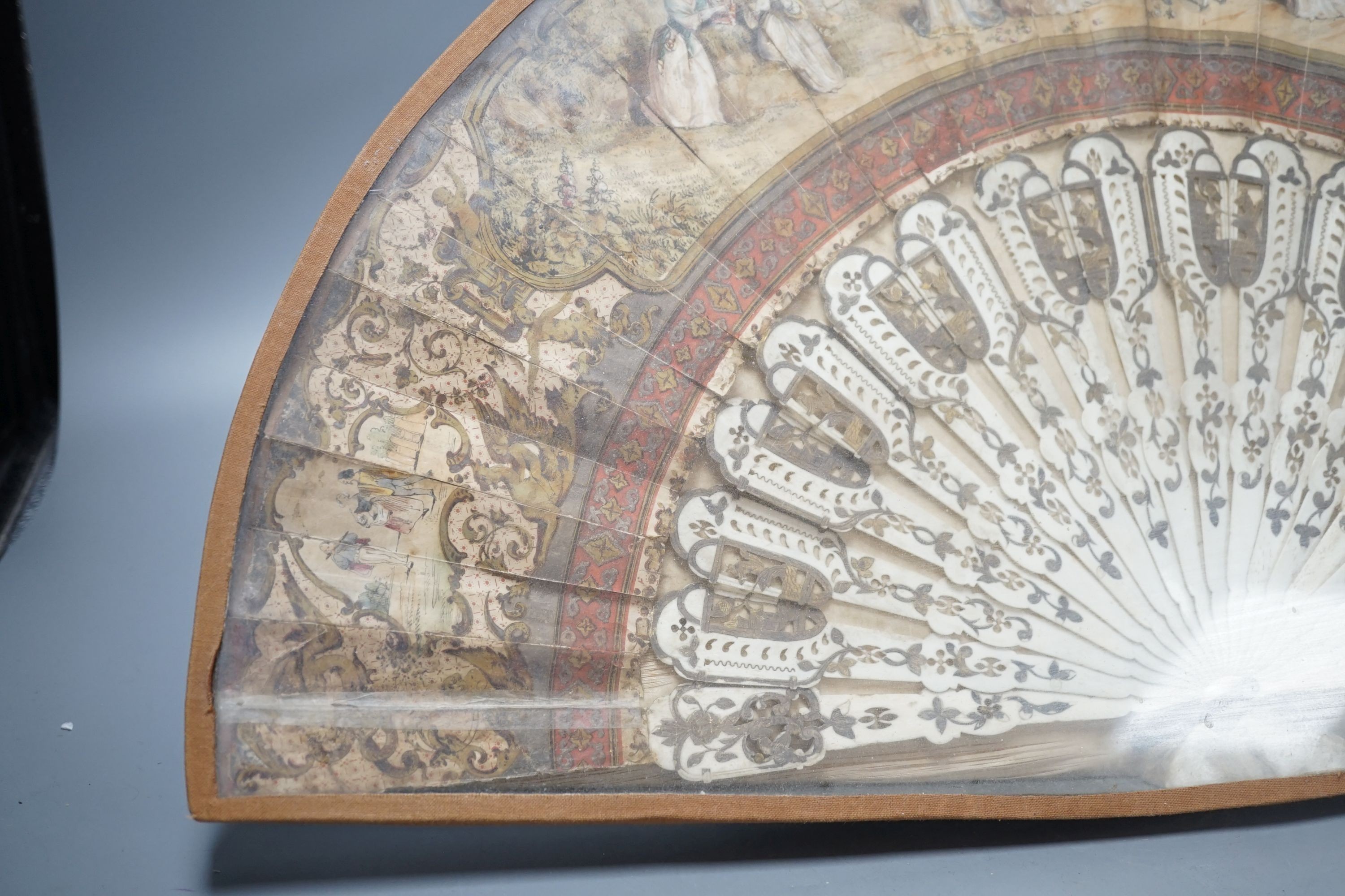 A cased 19th century French lithograph paper leaf and bone stick fan, 56 cms wide.